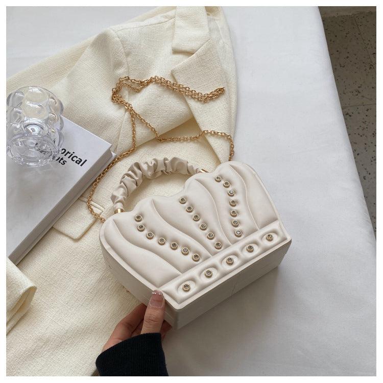 Elegant beige Crown Handbag with gold chain strap and stud detailing placed on a white fabric background.